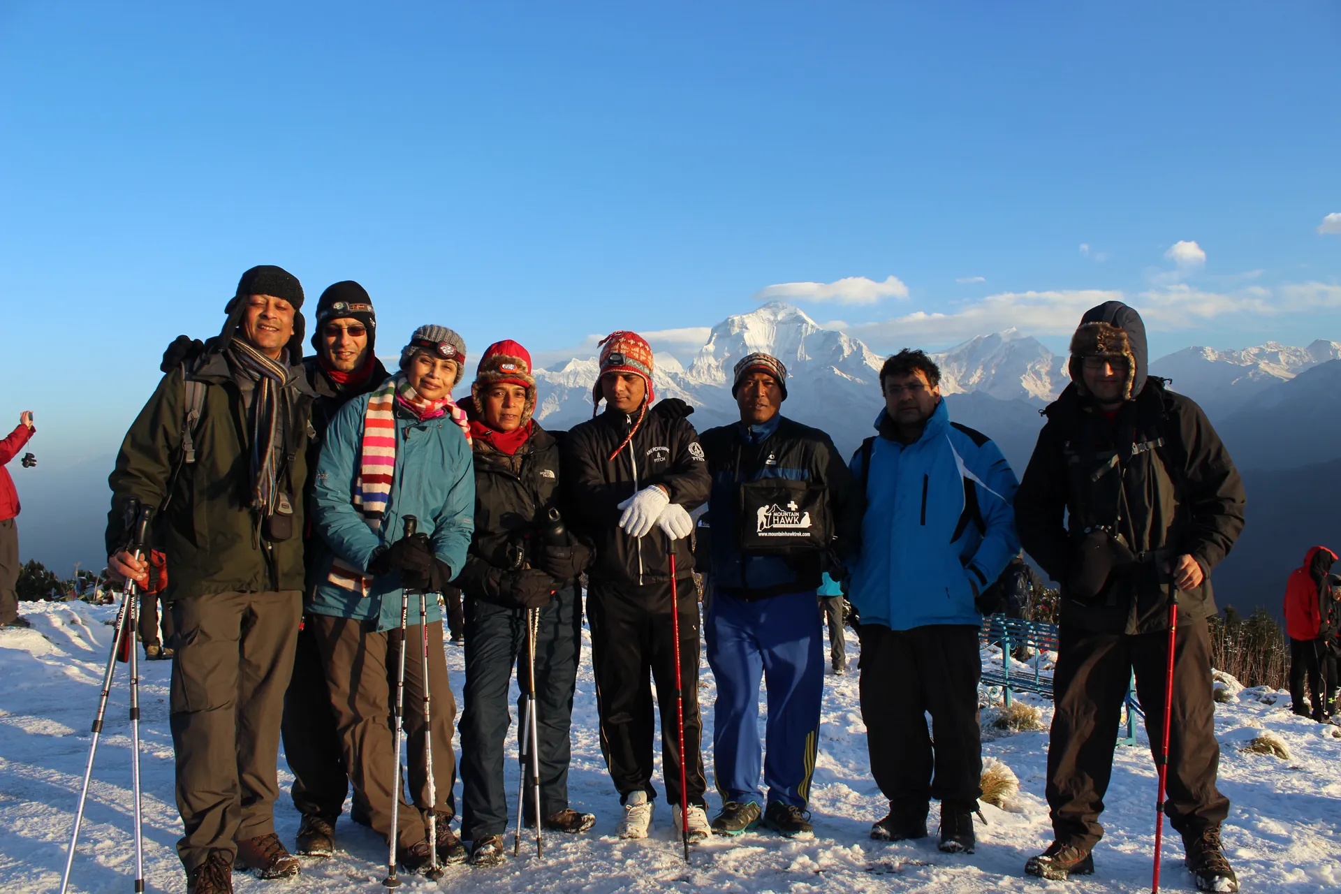 Eight trekkers posing on a snowy summit with Dhaulagiri visible in the distance