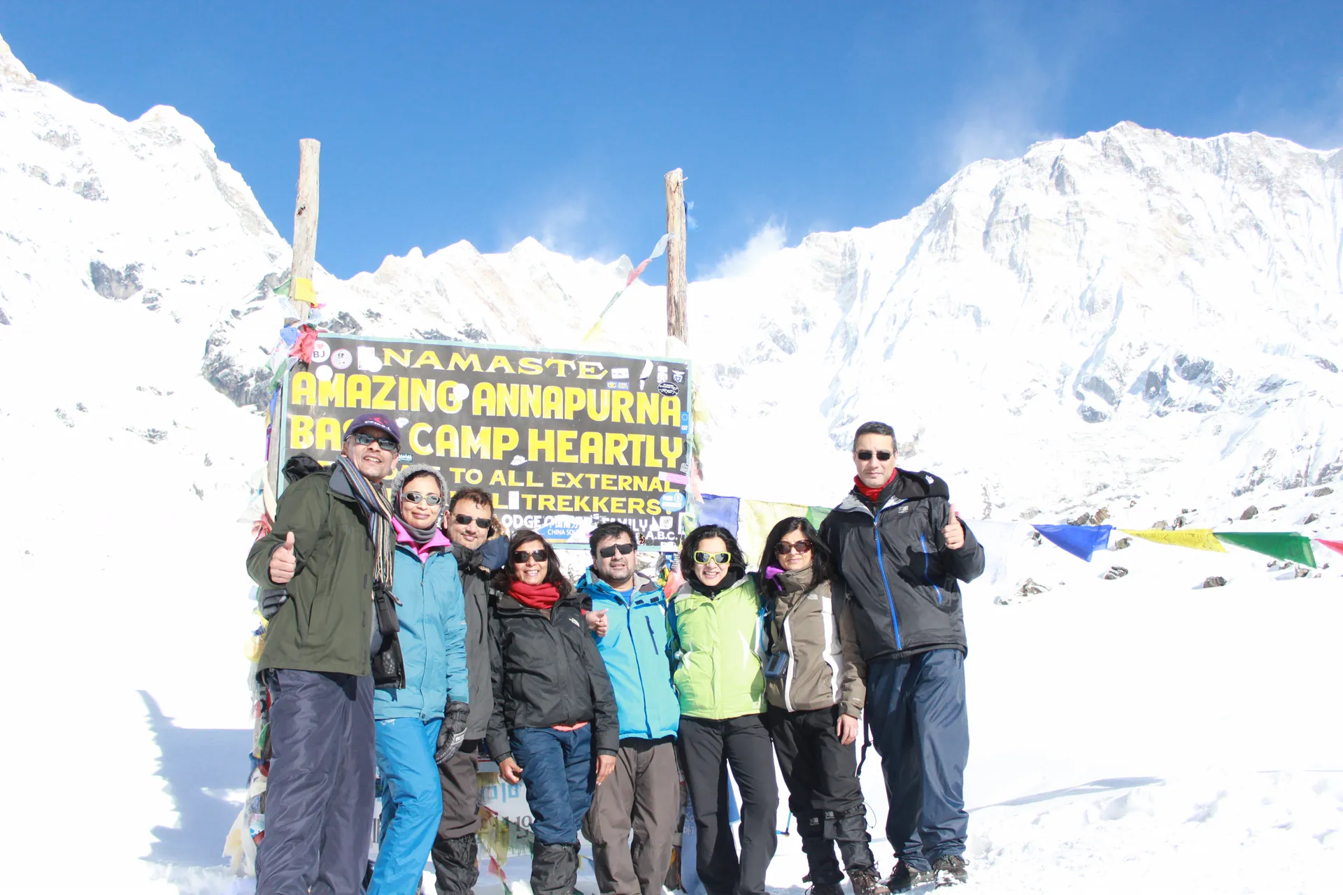 Seven trekkers at the snow-covered ABC sign with prayer flags and peaks behind