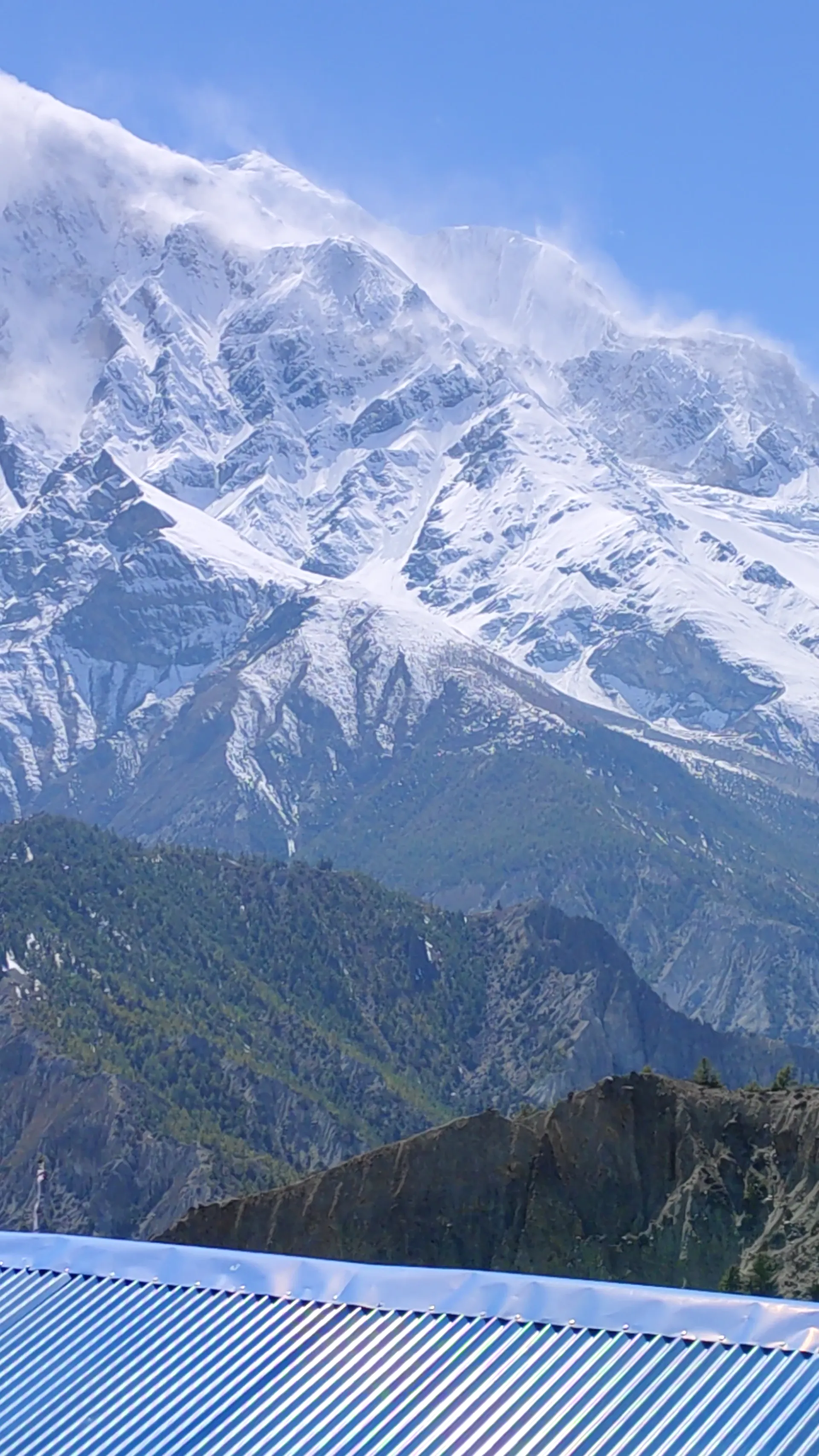 Snow-covered Annapurna peak with wind plume seen above a blue tin roof