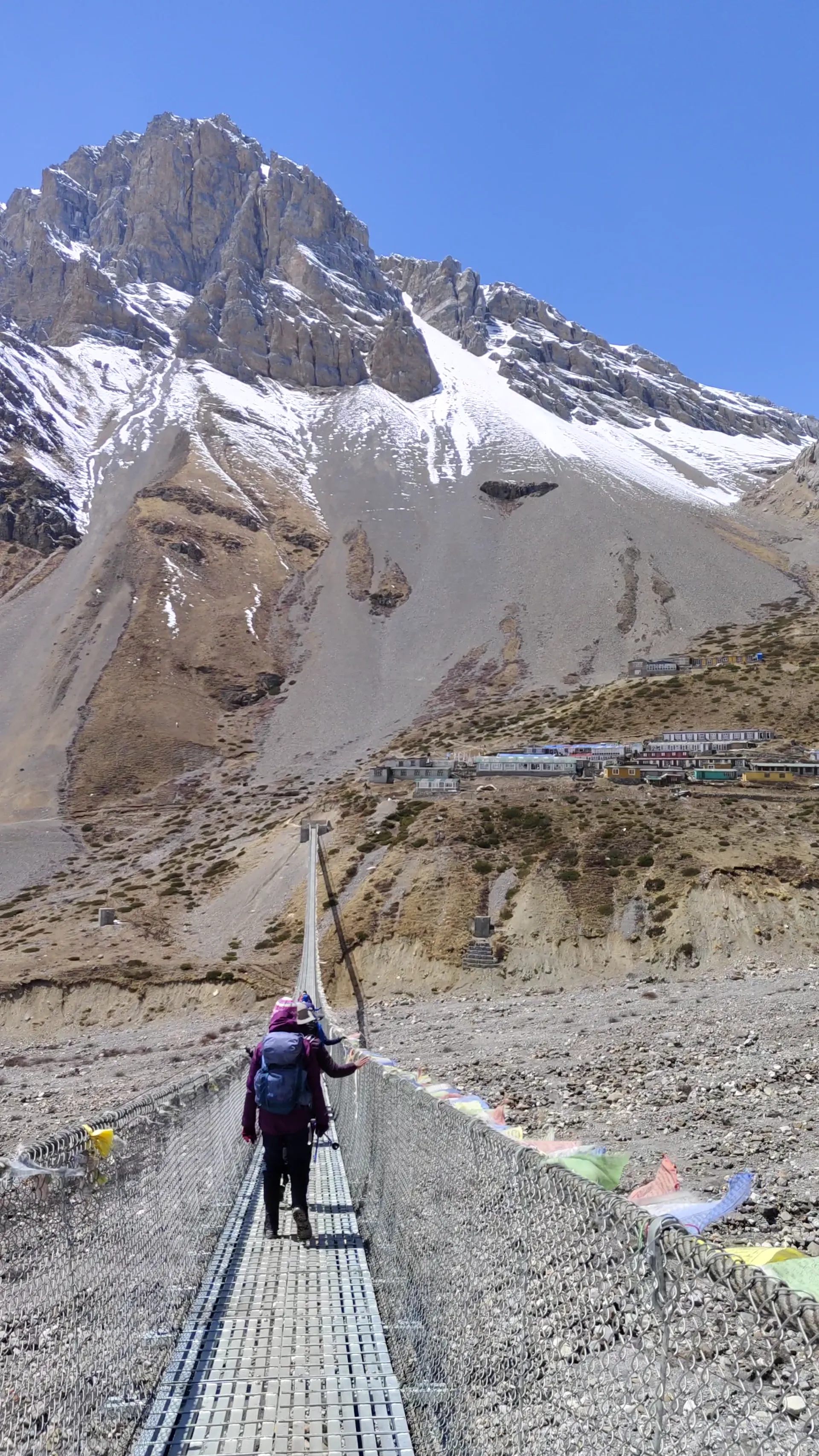Trekker crossing a metal bridge lined with prayer flags toward a rocky peak
