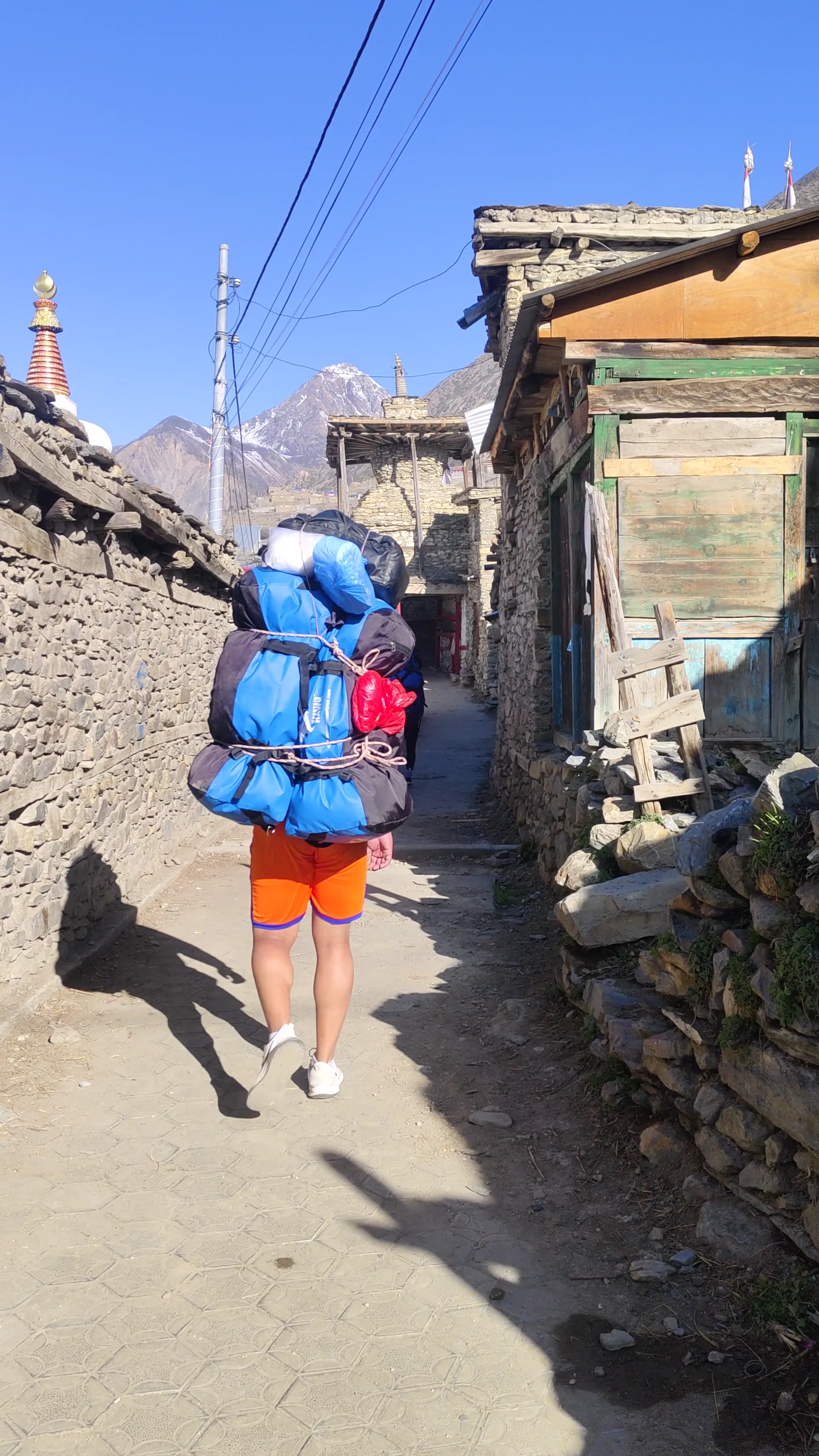 Porter carrying large blue bags through a narrow stone alley past a stupa