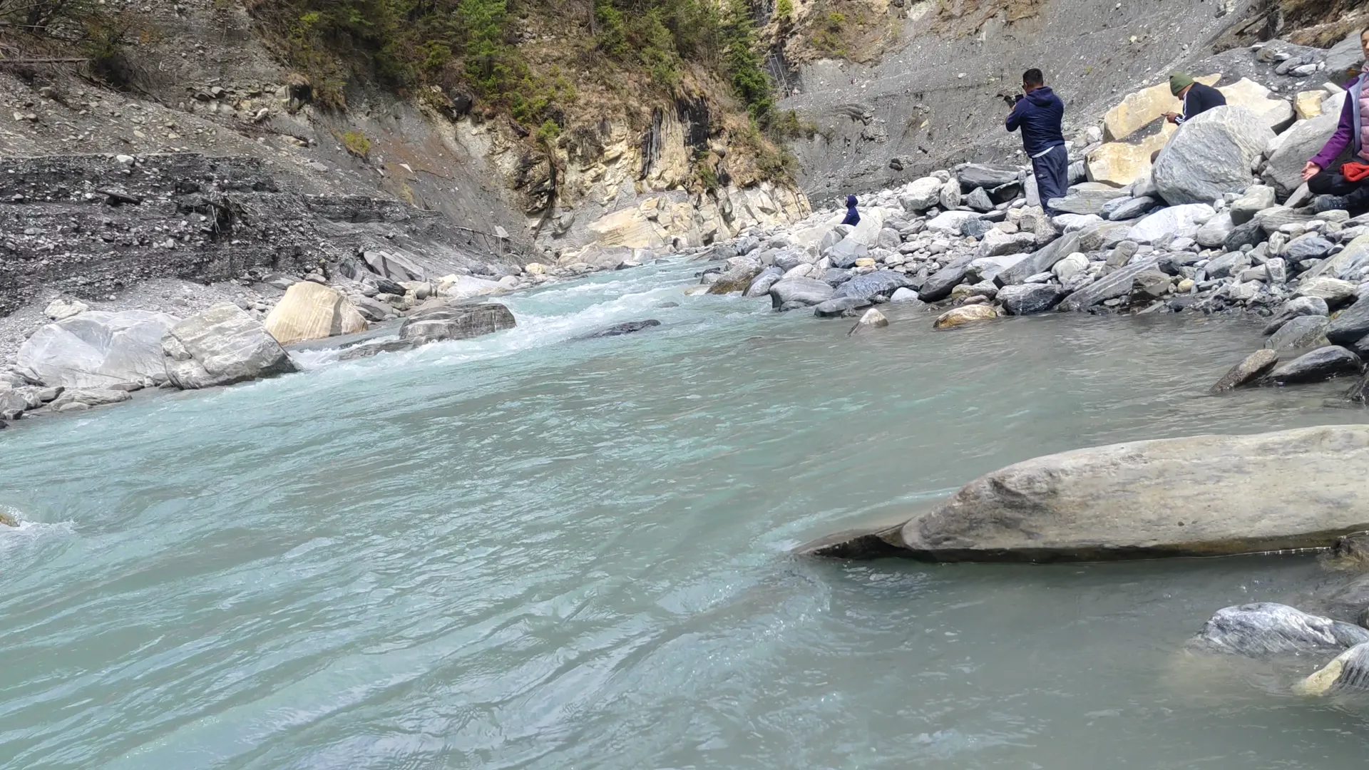 Trekkers standing on rocks beside a turquoise glacial river in a gorge