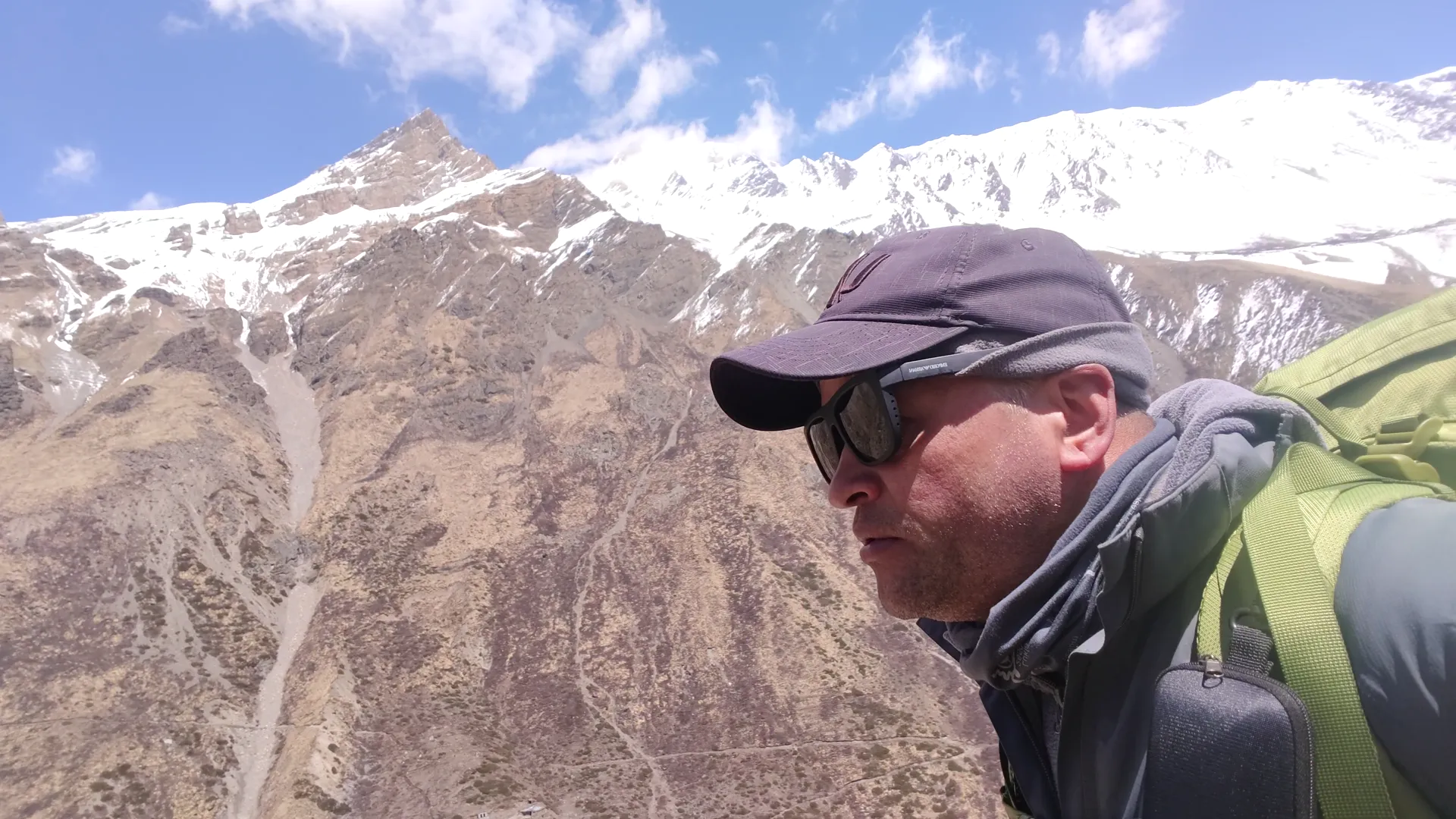Trekker selfie in cap and sunglasses with barren slopes and peaks behind