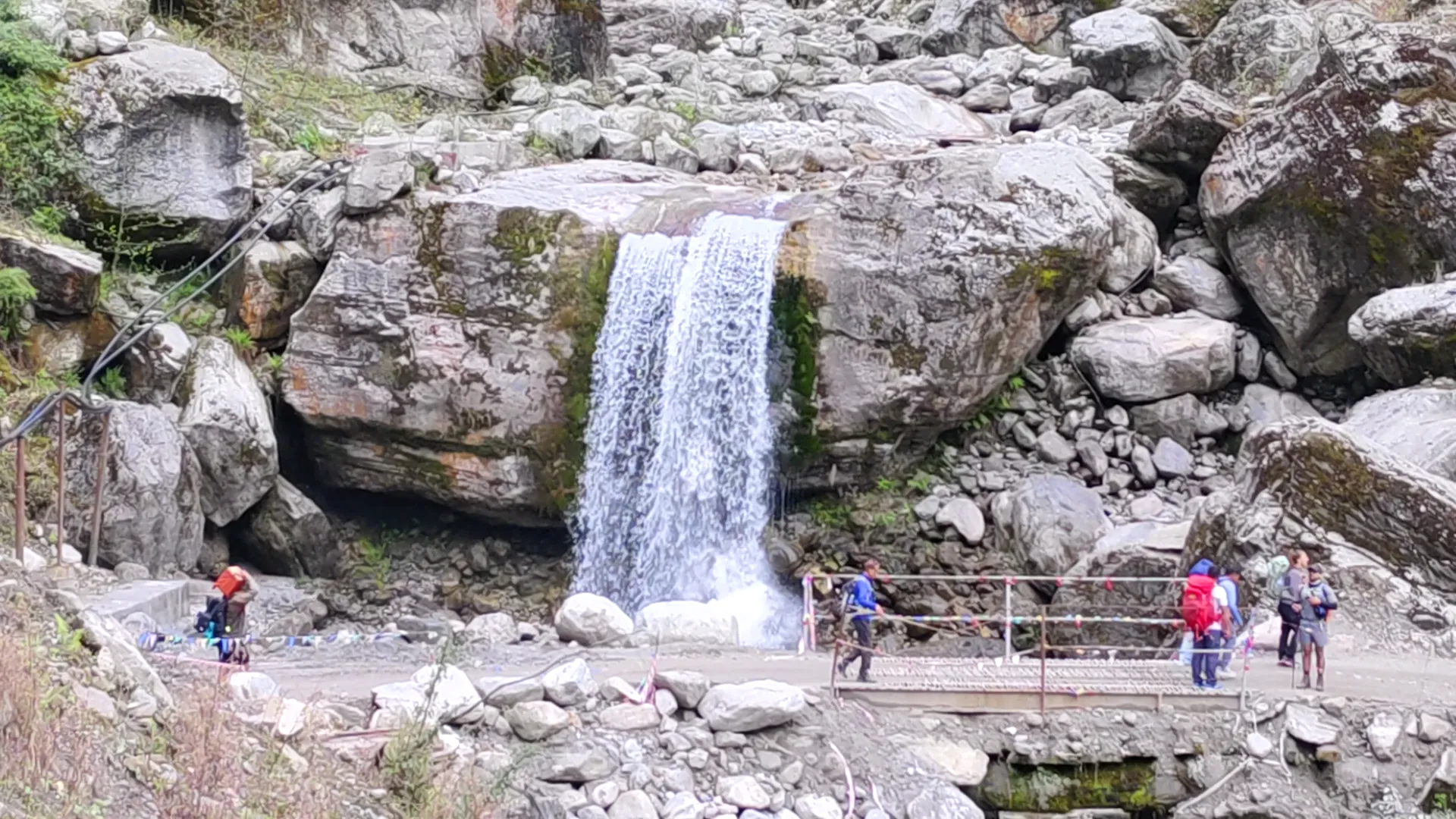 Trekkers on a metal footbridge below a waterfall cascading over boulders