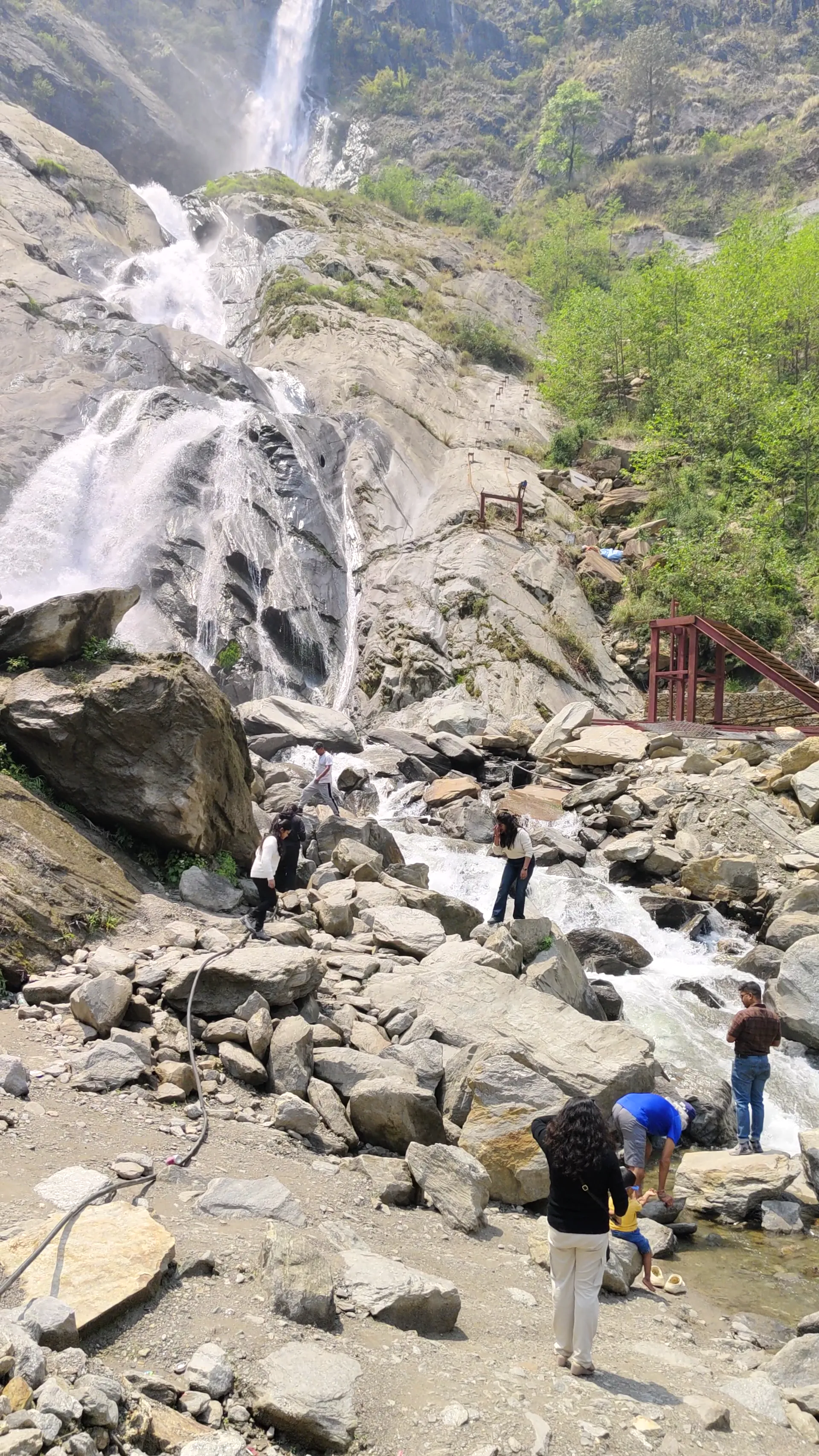 Trekkers scrambling on rocks near a tall waterfall with a red footbridge