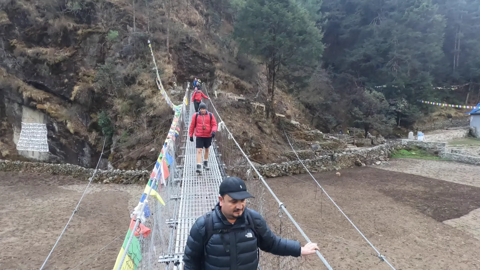 Prem crossing a suspension bridge with prayer flags and trekkers behind