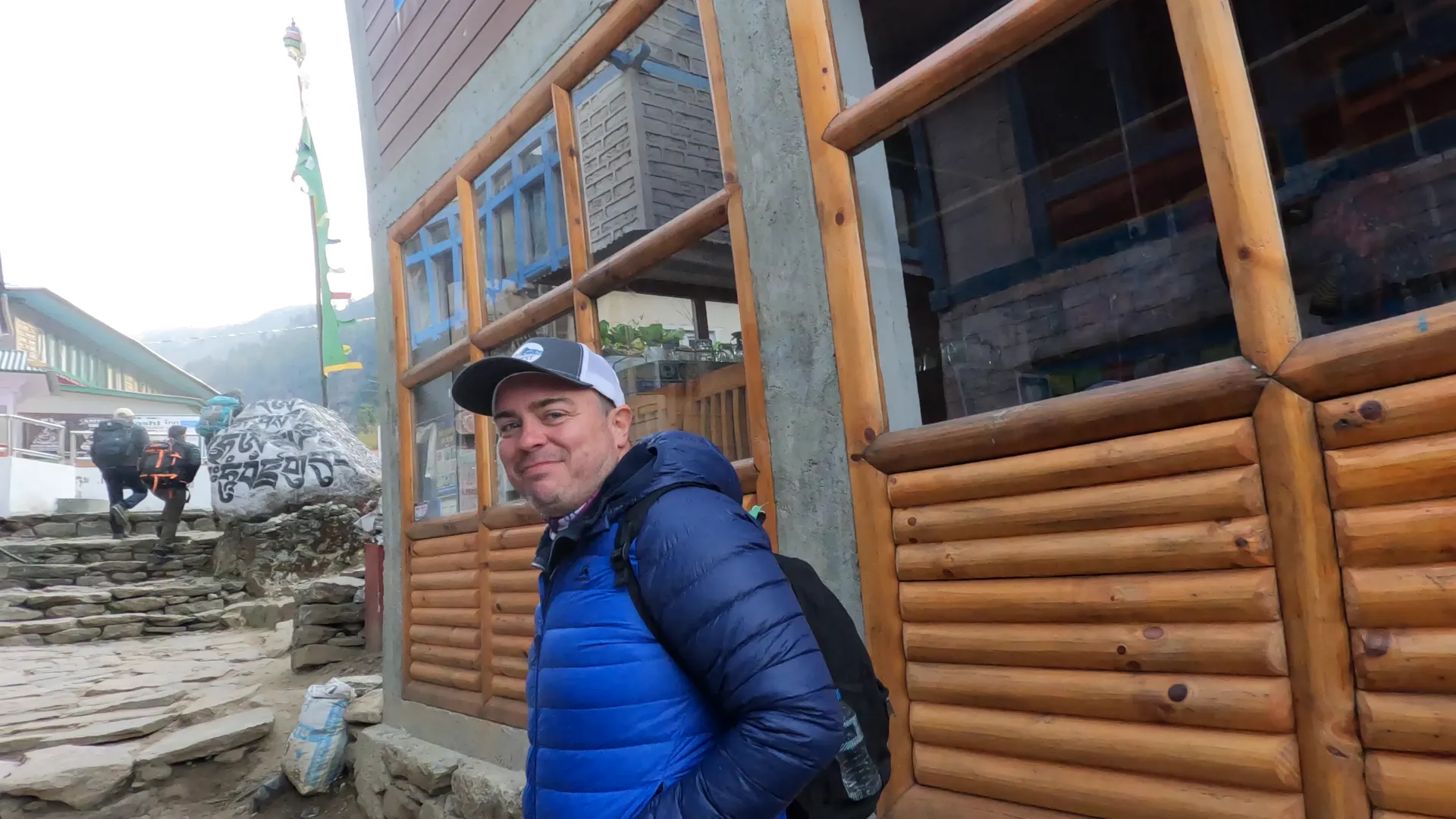 Trekker in blue jacket smiling outside a teahouse with mani stones