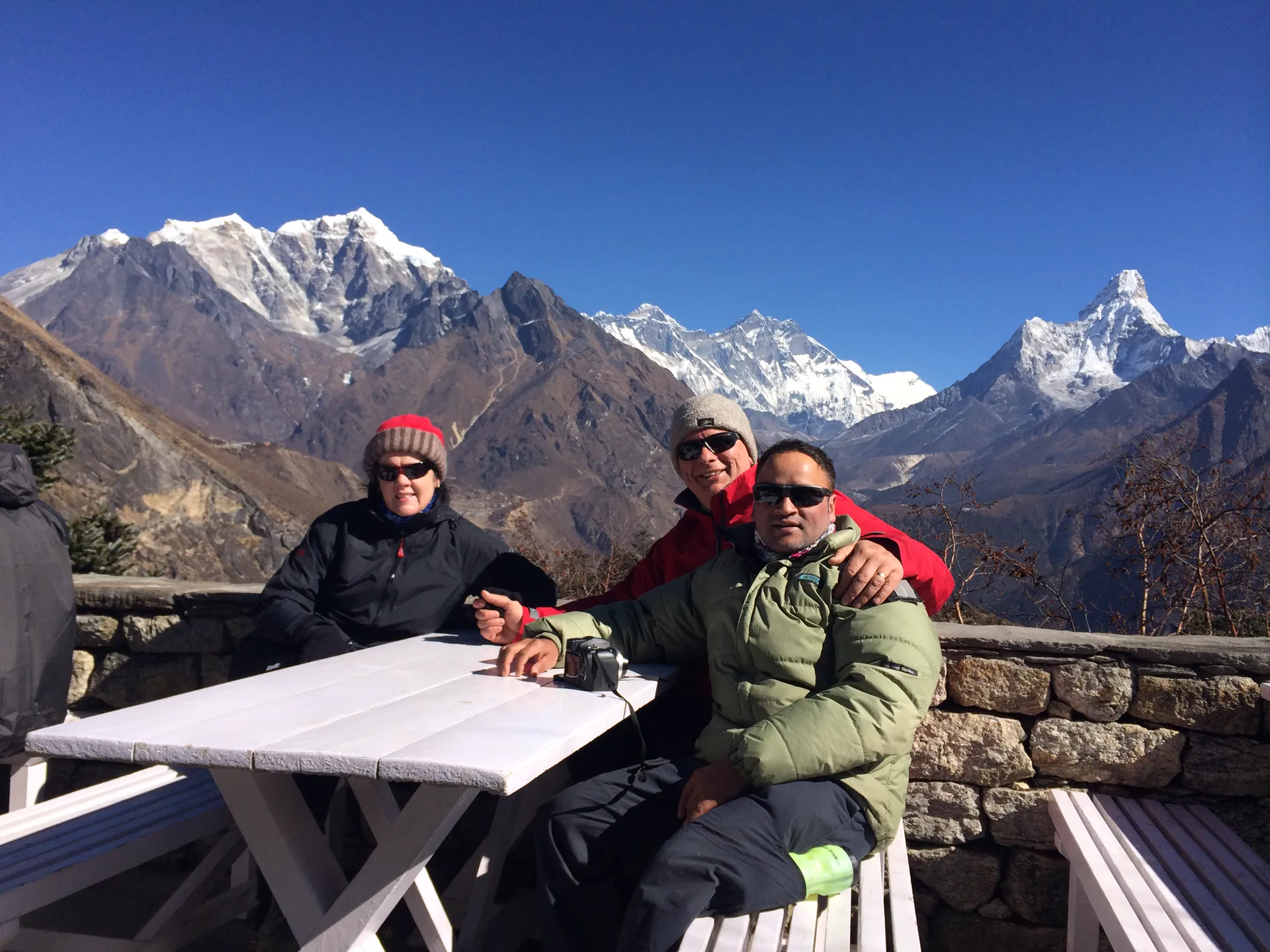Three trekkers at a teahouse with Everest, Lhotse, and Ama Dablam behind
