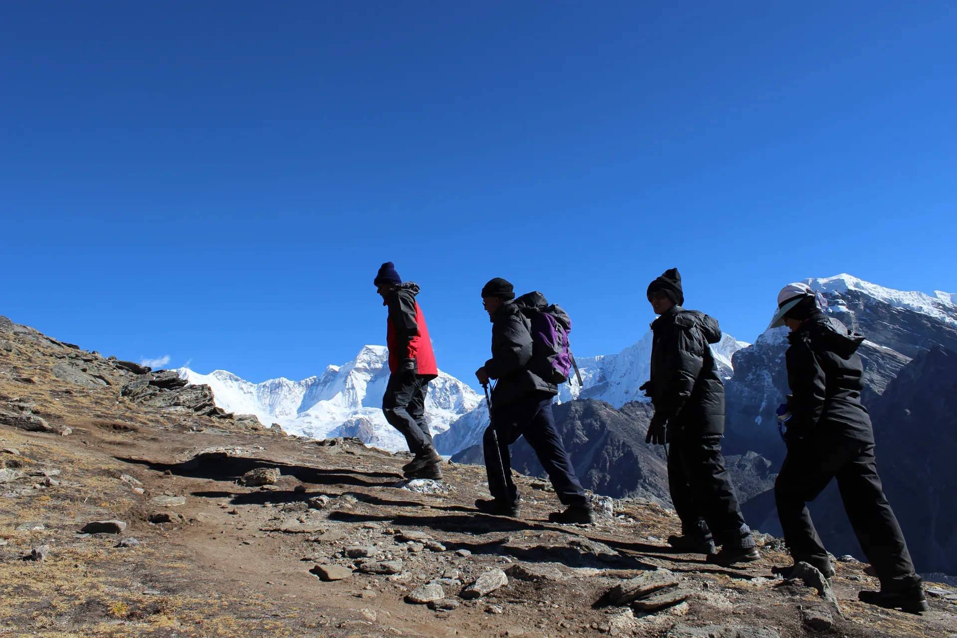 Five trekkers silhouetted walking single file on a ridge with snow peaks