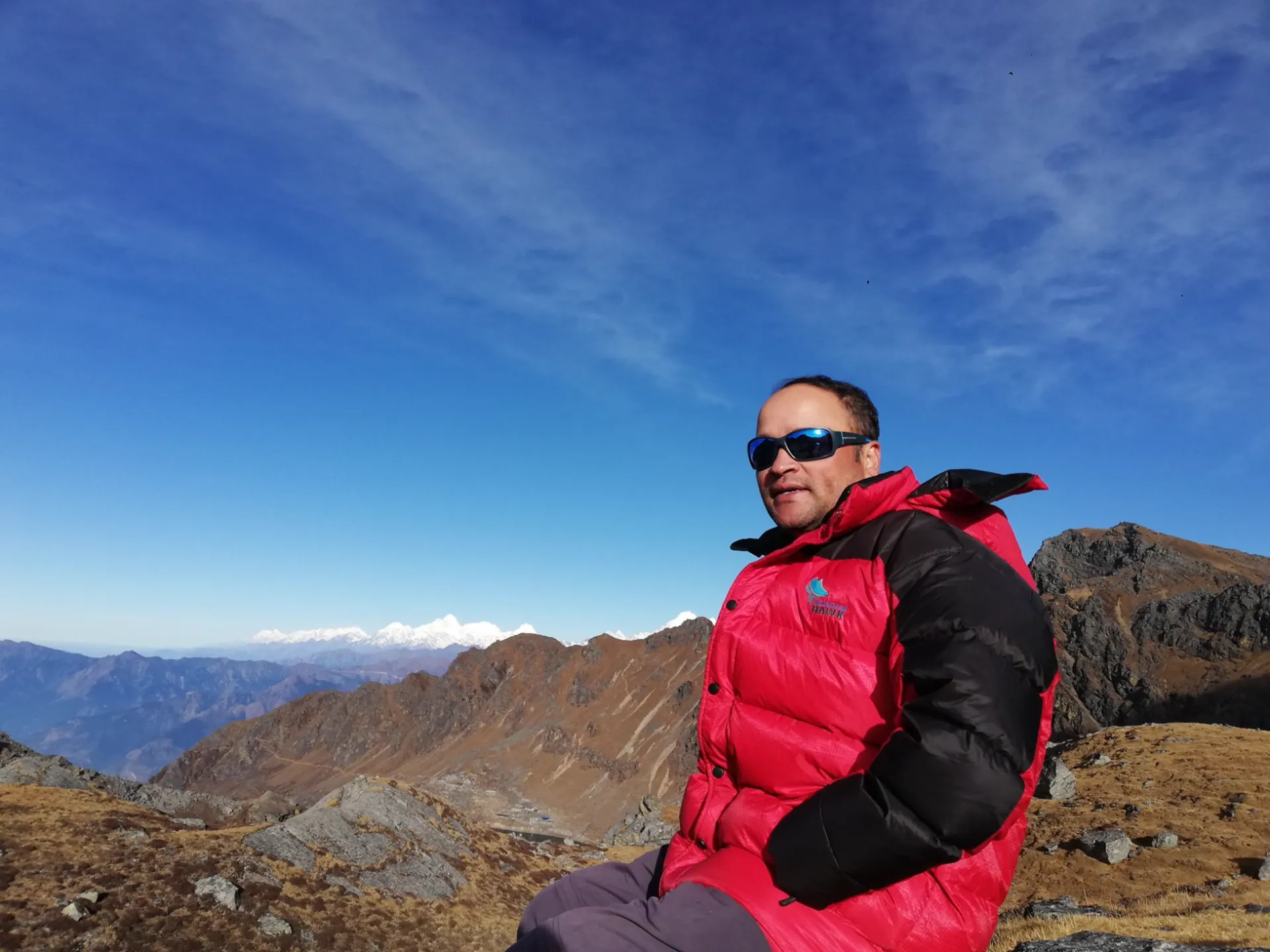 Guide in red down jacket and sunglasses resting at a high pass with ridges behind