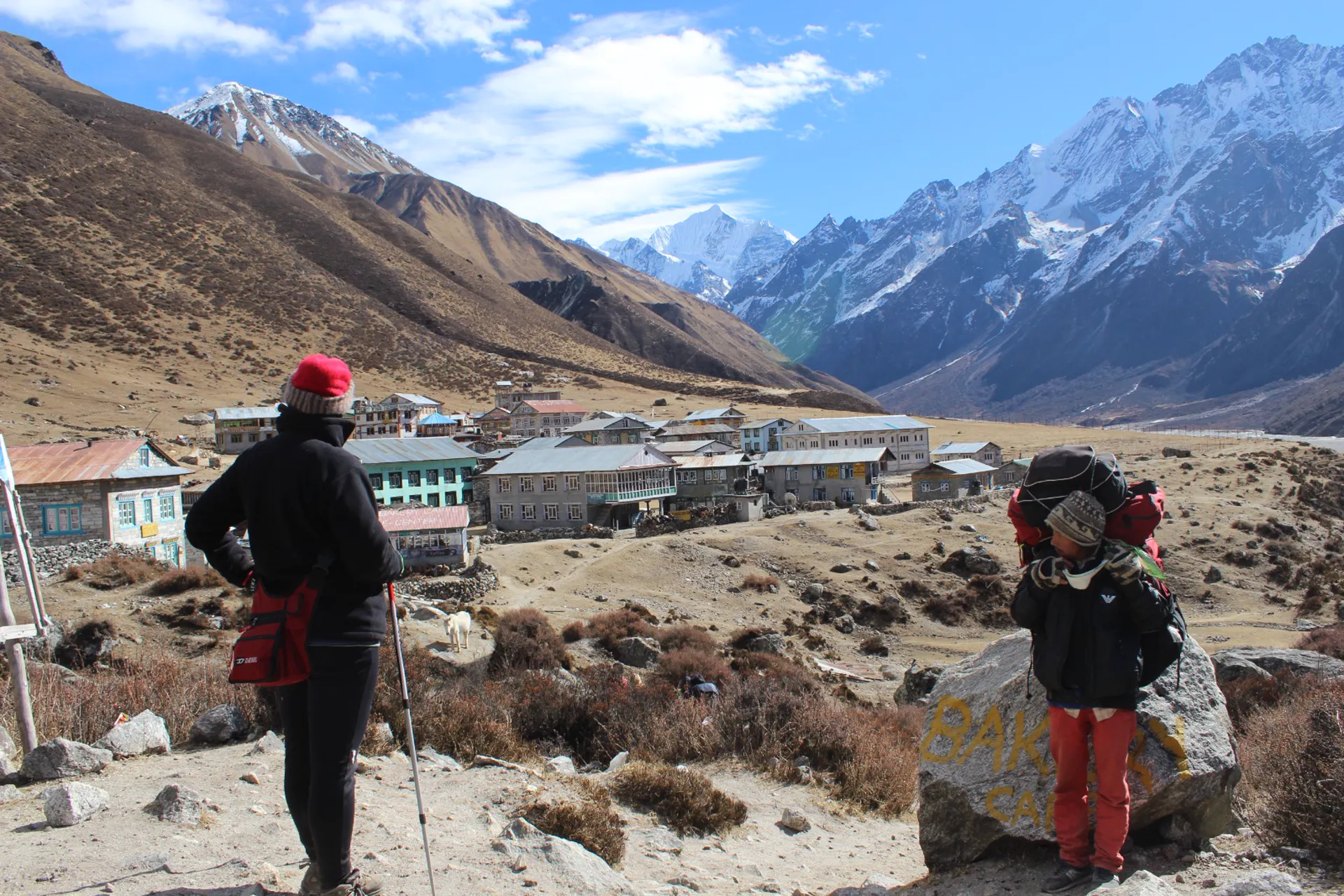Two trekkers and a porter with heavy load looking toward a mountain village