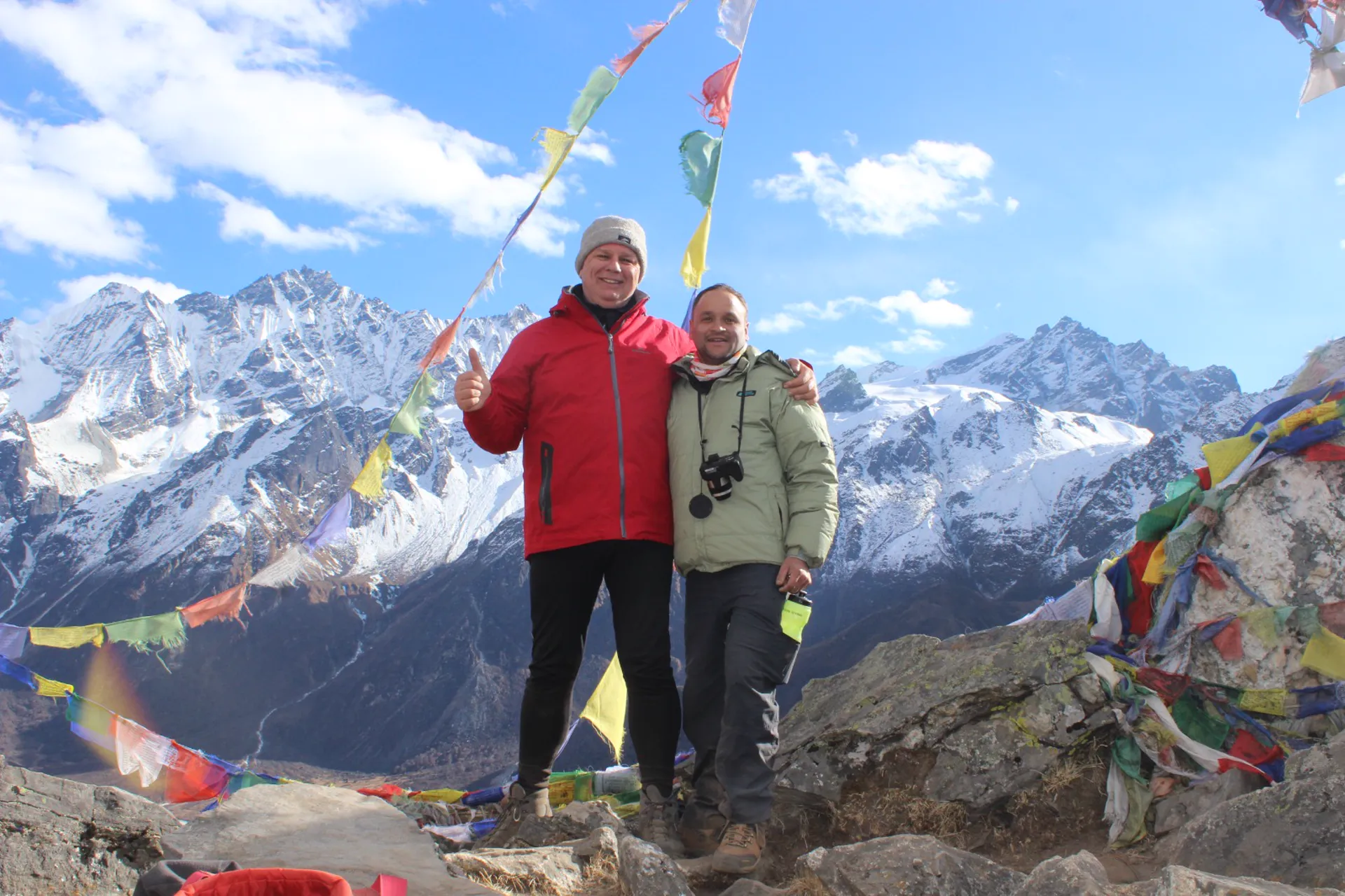 Trekker and guide with thumbs up among prayer flags at a viewpoint with glaciers