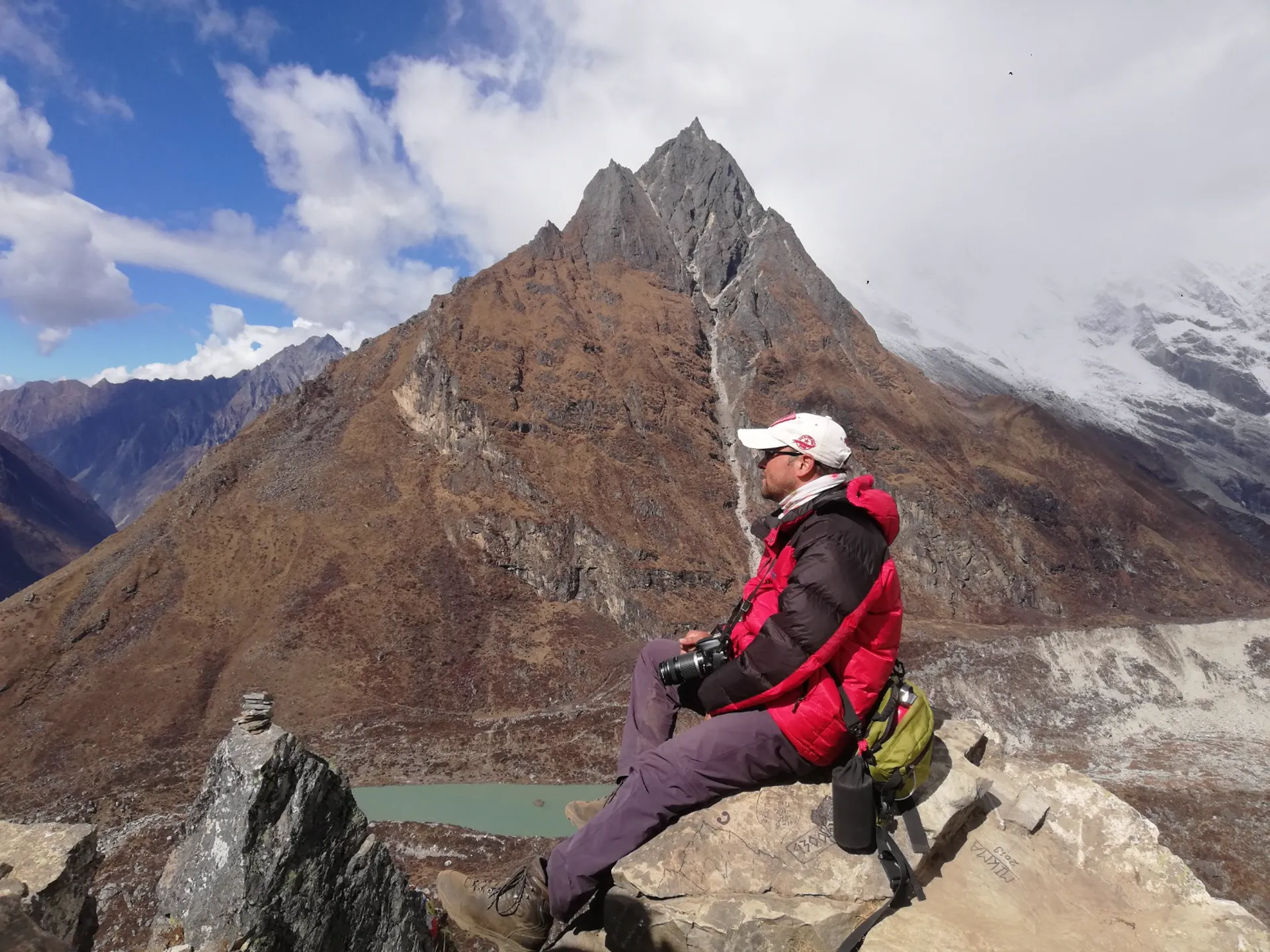 Guide sitting on a rock ledge with camera, sharp pyramid peak and glacial lake below