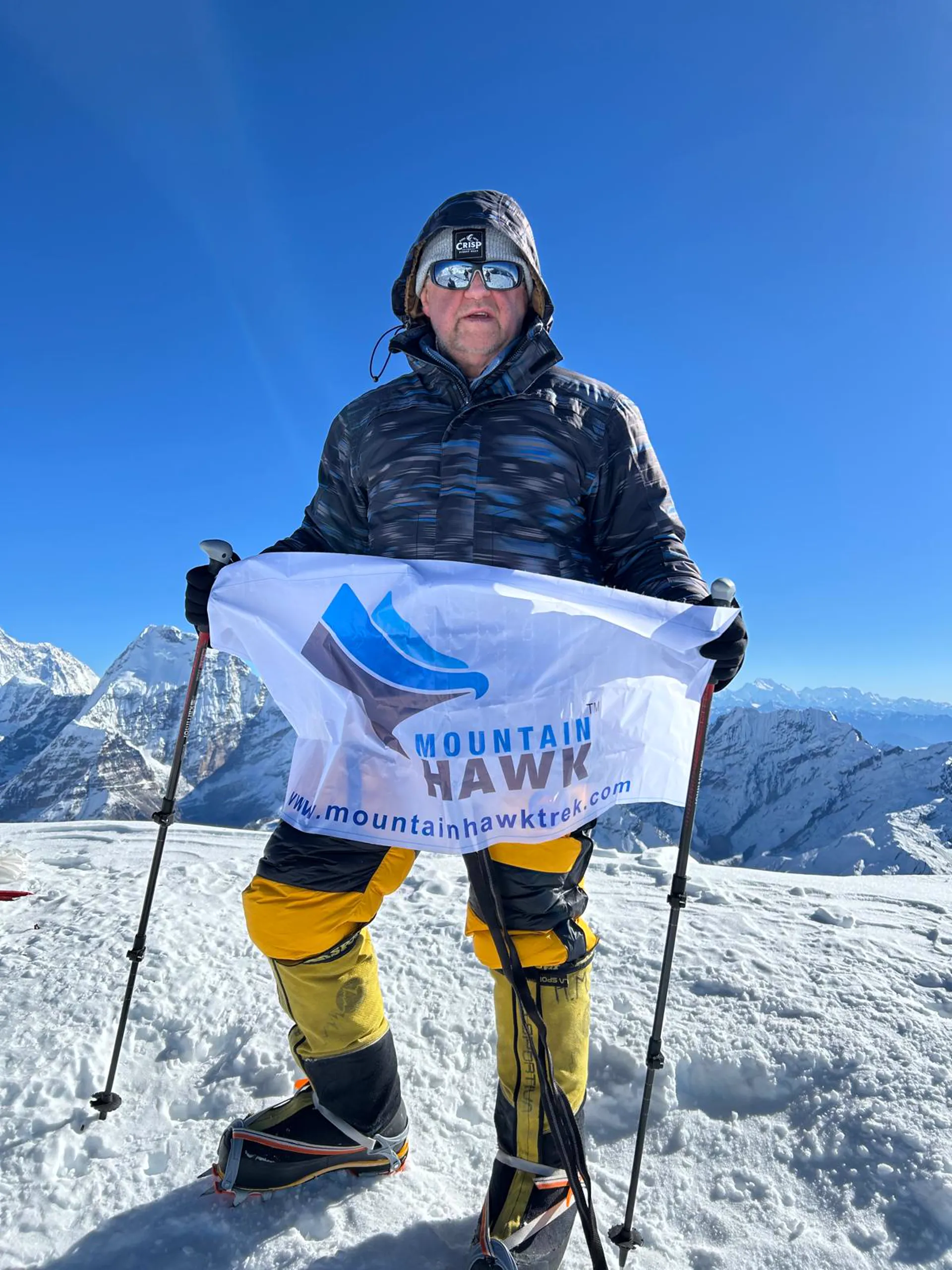 Climber holding Mountain Hawk Trek banner on snowy summit with Himalayan panorama