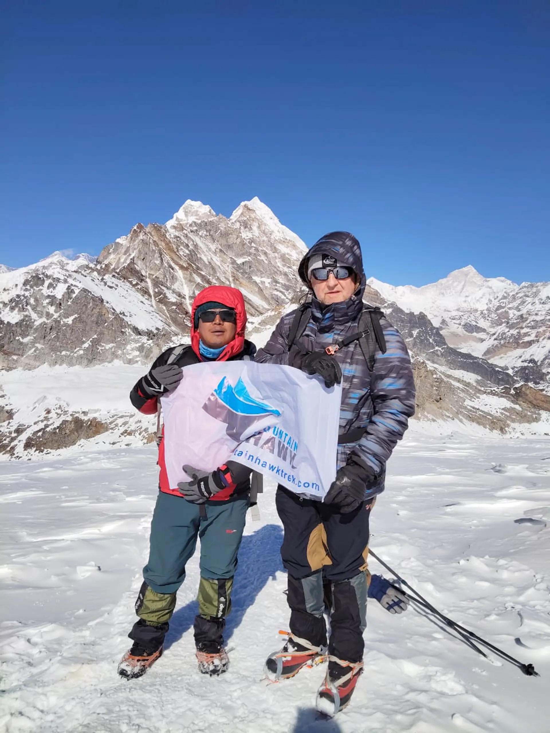 Guide and climber holding Mountain Hawk Trek banner on snow with peaks behind