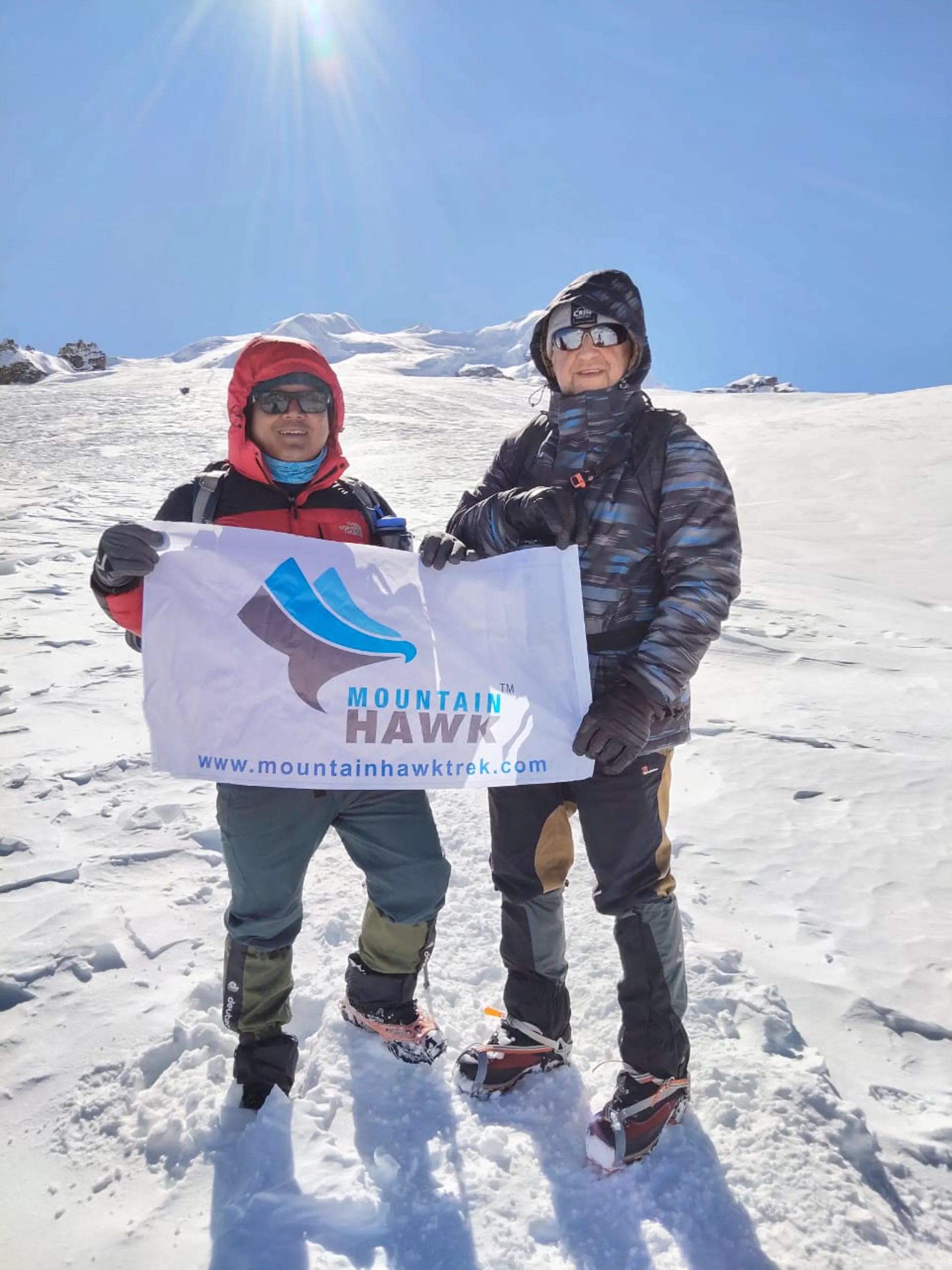 Guide and climber holding Mountain Hawk Trek banner on snowy slope with sun flare