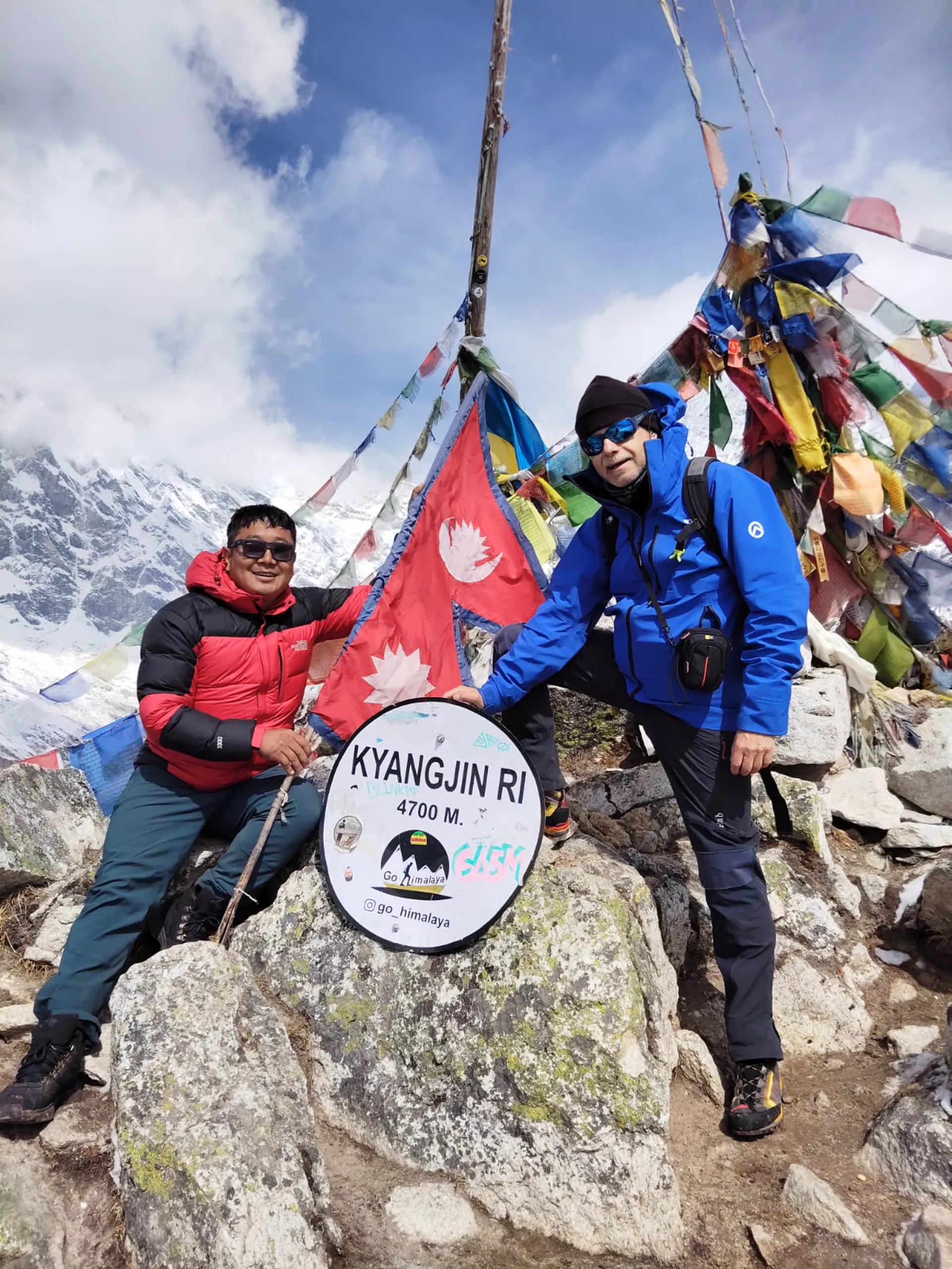 Two trekkers at Kyangjin Ri 4,700m sign with Nepal flag and prayer flags