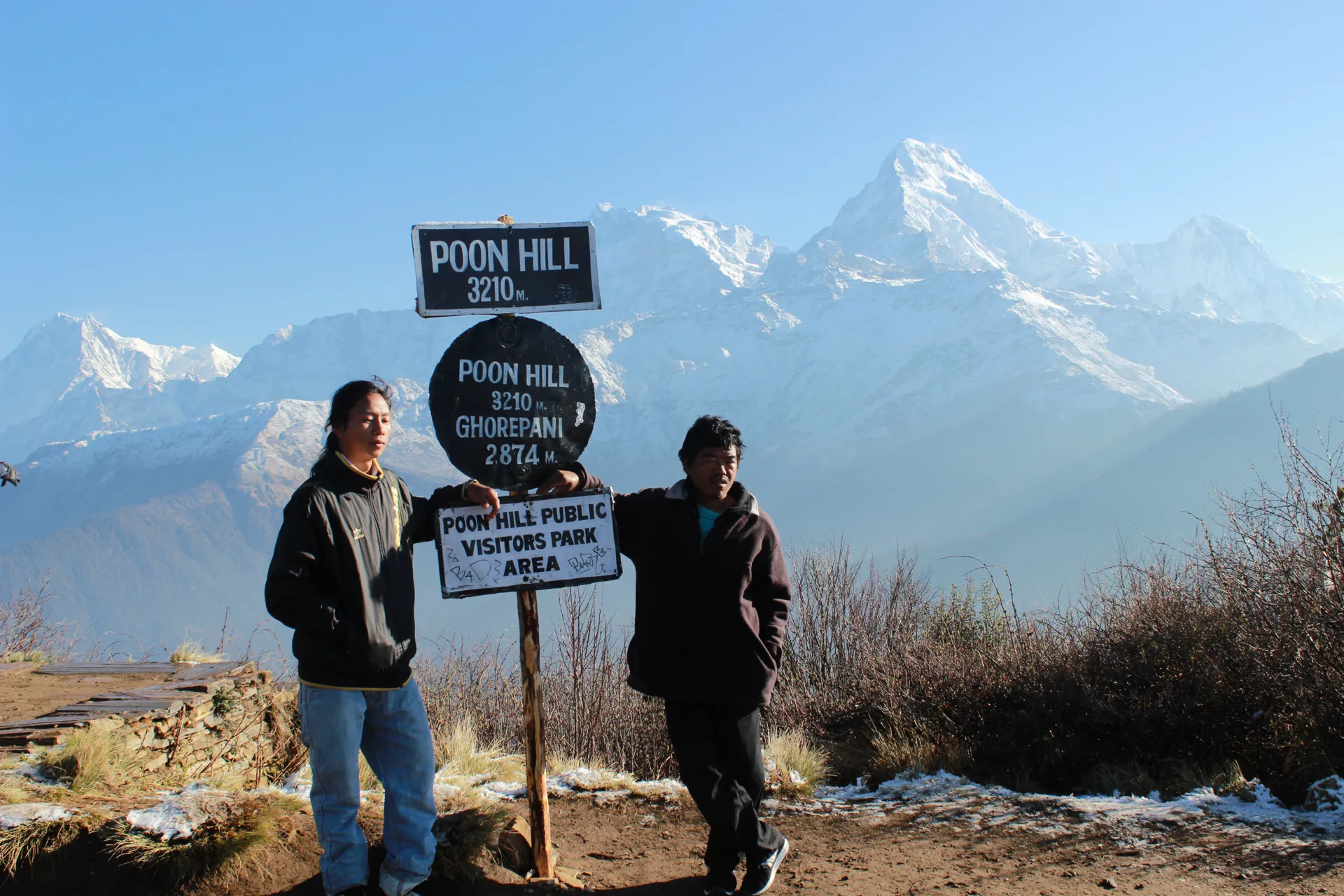 Two trekkers at the Poon Hill and Ghorepani signs with snowy peaks behind