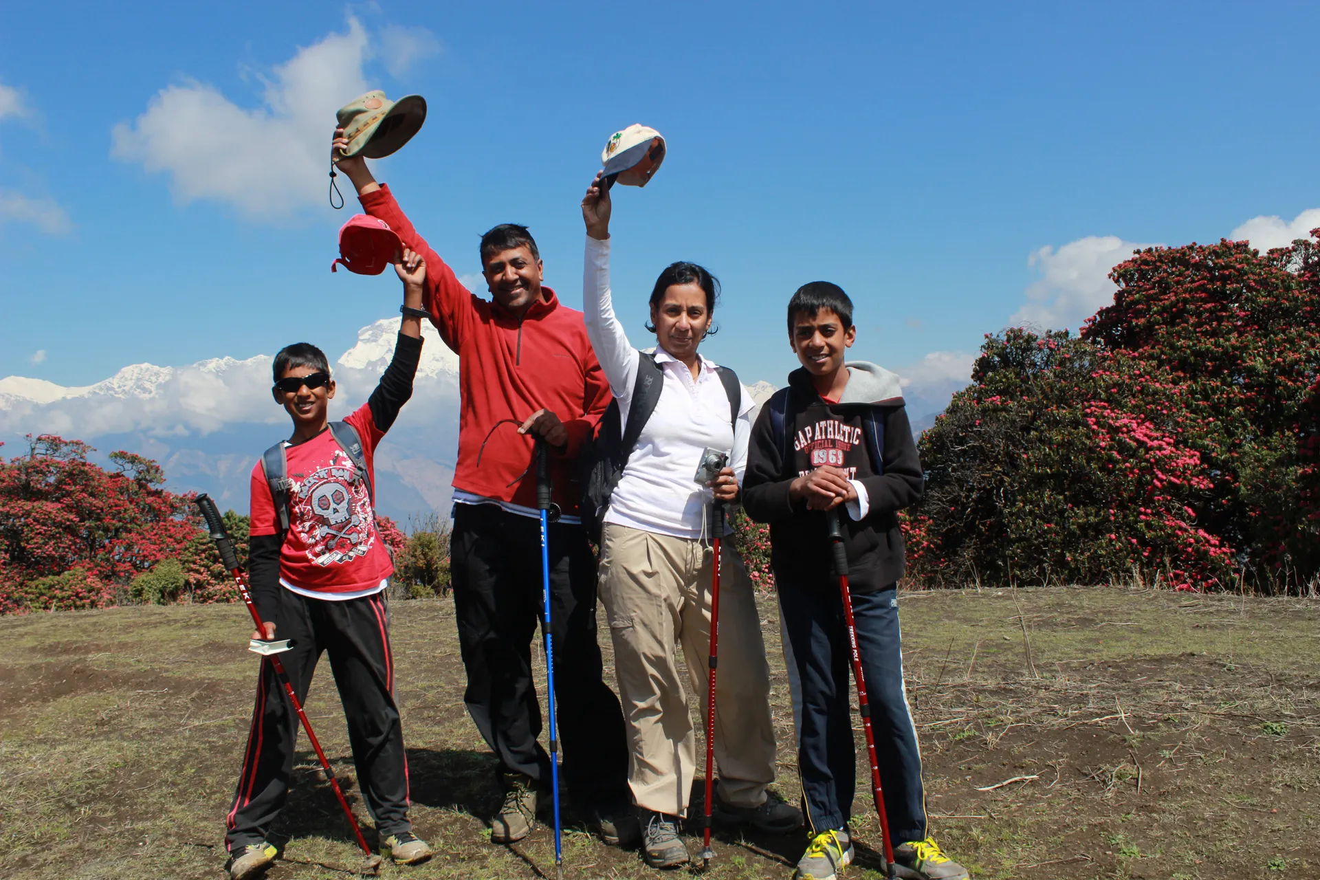 Family of four waving hats on a hilltop with rhododendrons and mountains